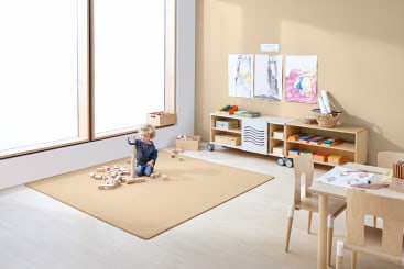 Ein kleines Kind sitzt auf einem beigen Teppich und spielt mit Holzklötzen in einem Klassenzimmer mit hellen Holzmöbeln, Kunstzubehör und Kinderzeichnungen an der Wand. Große Fenster sorgen für natürliches Licht.