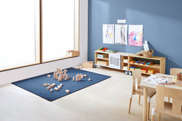 In einem Klassenzimmer mit hellen Holzmöbeln stehen ein Tisch mit Malutensilien, ein Teppich mit Holzklötzen, Regale mit Körben und Spielzeug sowie Kinderkunstwerke an einer blauen Wand neben einem großen Fenster.