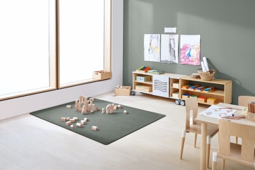 Ein aufgeräumtes Klassenzimmer mit einem kleinen Tisch und Stühlen, Regalen mit Malutensilien, Kinderzeichnungen an der Wand und Holzklötzen, die auf einem Teppich in der Mitte des Raums verstreut sind. Durch ein großes Fenster fällt Licht ins Zimmer.