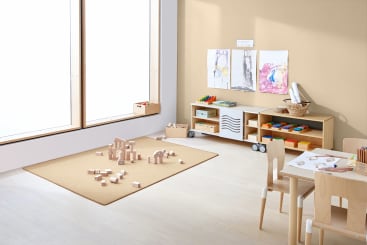 Ein helles Klassenzimmer mit Holzmöbeln, einem Tisch mit Malutensilien, Regalen mit Körben und Behältern, Kinderzeichnungen an der Wand und Holzklötzen, die auf einem beigen Teppich in der Nähe eines großen Fensters verstreut sind.