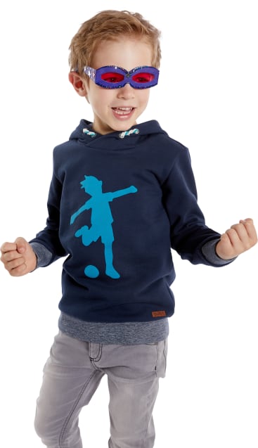 Ein kleiner Junge trägt ein blaues Sweatshirt mit einer Fußballergrafik, eine graue Hose und eine rot-blaue maskenartige Brille. Er lächelt und hält seine Fäuste in einer aufgeregten Pose hoch.