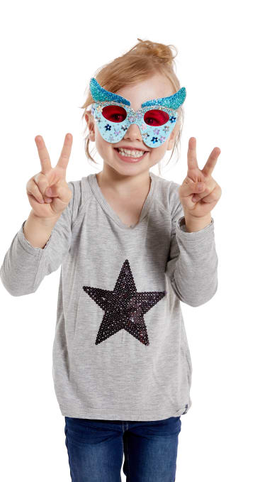Ein kleines Kind, das ein graues Hemd mit einem Paillettenstern, blaue Jeans und eine dekorative blau-rote Brille trägt, steht lächelnd vor einem weißen Hintergrund und macht mit beiden Händen Friedenszeichen.