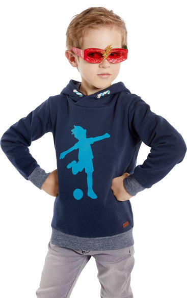 Ein Junge mit einer roten Glitzerbrille und einem marineblauen Kapuzenpulli mit einem hellblauen Fußballspielermotiv steht mit den Händen in den Hüften und schaut zur Seite.