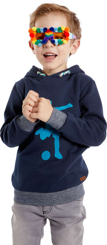 Ein kleines Kind steht lächelnd da und trägt einen marineblauen Kapuzenpullover und eine hellgraue Hose. Das Kind hat bunte Bommeln an seiner Brille befestigt, die seine Augen verdecken. Der Hintergrund ist einfarbig weiß.