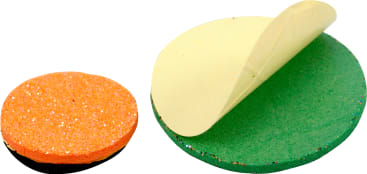 Zwei runde Schaumstoffkreise sind abgebildet; der linke ist orange und mit Glitter überzogen, während der rechte grün ist und eine gelbe Klebefläche hat, die teilweise abgezogen wurde.