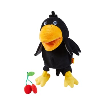 Eine schwarze Plüschkrähe mit gelben Füßen und Schnabel steht aufrecht neben einer kleinen Stoffkirsche mit zwei roten Kirschen und einem grünen Blatt, alles auf weißem Hintergrund.