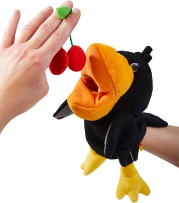 Eine Hand hält zwei rote Pom-Pom-Kirschen mit einem grünen Filzblatt vor einer schwarz-gelben Vogelpuppe mit offenem Maul, die von einer anderen Hand gesteuert wird, alles vor einem weißen Hintergrund.