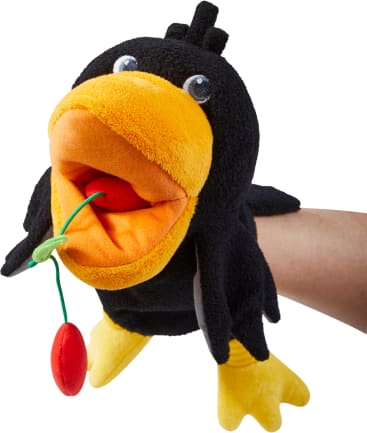 Eine schwarz-gelbe Vogelhandpuppe mit weit geöffnetem Schnabel hält ein rotes und grünes Stoffobjekt in ihrem Mund. In der Puppe, die vor einem weißen Hintergrund fotografiert wurde, befindet sich ein Personenarm.