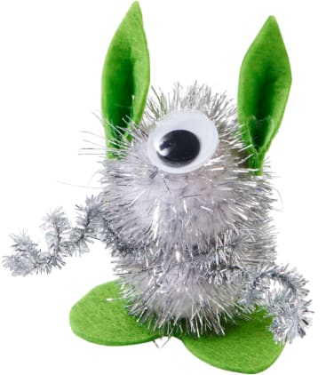 Ein kleines Spielzeugwesen aus silbernem Lametta mit einem großen Glotzauge, grünen Filzohren und grünen Filzfüßen, das aufrecht sitzt und einen gebogenen Lamettaarm hält.