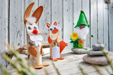 Drei handgefertigte Kunstwerke aus Holz, Filz und anderen Materialien sind auf einer Holzfläche ausgestellt. Sie stellen einen Hasen, einen Fuchs und einen Gnom mit grüner Mütze dar, die vor einem verwitterten Holzhintergrund stehen.