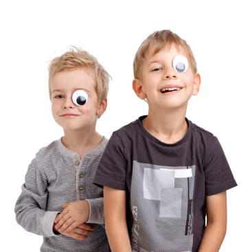 Zwei kleine Jungen stehen nebeneinander und lächeln in die Kamera. Jeder Junge hat einen großen Plastikaufkleber mit Glupschaugen auf einem seiner Augen. Der Hintergrund ist schlicht und weiß.