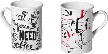 Abgebildet sind zwei weiße Keramikbecher. Die linke Tasse hat die Phrase all you need is coffee in schwarzer Zierschrift mit einer kleinen Kaffeetassen-Illustration. Die rechte Tasse zeigt ein abstraktes Design in schwarzer und roter Farbe.