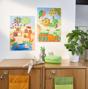 Zwei bunte, kindliche Kunstposter hängen an einer weißen Wand über einem hellen Holzschrank. Auf dem Schrank stehen ein Plüschkaninchen, eine grüne Brotdose, eine Metallflasche und eine Topfpflanze mit gefleckten Blättern.