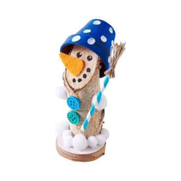 Eine handgefertigte Schneemannfigur aus einem senkrechten Holzstamm, mit einer blau gepunkteten Tasse als Hut, Knopfverzierungen, weißen Bommeln, einer Karottennase aus Filz und einem gestreiften Strohhalm, der einen kleinen Besen hält.