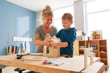 Eine Frau und ein Junge arbeiten gemeinsam an einem Holzprojekt an einem Tisch in einem hellen Raum. Auf dem Tisch liegen verschiedene Werkzeuge und Holzteile, und im Hintergrund sind Regale mit Material zu sehen.