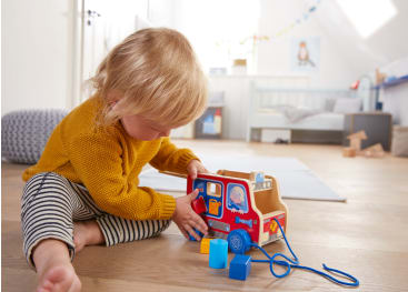 Ein kleines Kind in einem gelben Pullover sitzt auf einem Holzboden und spielt mit einem bunten Holzspielzeug, zu dem auch Formblöcke gehören, in einem hellen Raum mit hellem Dekor und einigen Spielzeugen im Hintergrund.