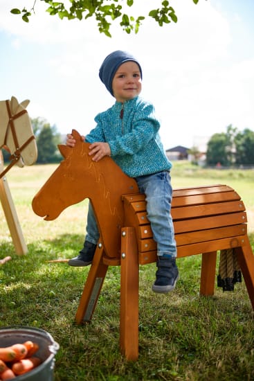 Ein kleines Kind in einem blauen Pullover, Jeans und einem blauen Hut sitzt auf einem hölzernen Spielzeugpferd im Freien im Gras. Im Hintergrund sind ein Eimer mit Spielzeugmöhren und ein weiteres Holzpferd zu sehen.