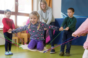 Ein junges Mädchen sitzt auf einer Holzschaukel im Haus und hält sich an Seilen fest, während andere Kinder und ein Erwachsener helfen. Alle tragen Freizeitkleidung, und der Raum ist durch ein großes Fenster hell erleuchtet.