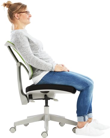 Eine Frau mit Brille, grauem Pullover, blauen Jeans und weißen Turnschuhen sitzt in einem modernen Bürostuhl mit Netzrücken, lehnt sich leicht zurück und lächelt vor einem weißen Hintergrund.