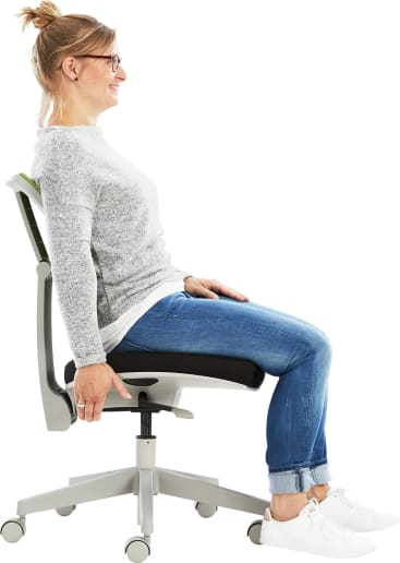 Eine Frau sitzt aufrecht auf einem Bürostuhl mit geradem Rücken, die Hände ruhen auf den Seiten des Sitzes, die Füße sind flach auf dem Boden. Sie trägt eine Brille, einen grauen Pullover, blaue Jeans und weiße Turnschuhe.