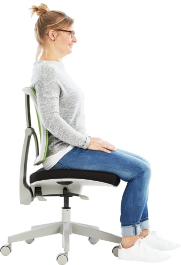 Eine Frau mit blondem Haar in einem Dutt, einer Brille, einem grauen Pullover, blauen Jeans und weißen Turnschuhen sitzt aufrecht auf einem Bürostuhl, die Füße flach auf dem Boden, die Hände auf die Oberschenkel gestützt.