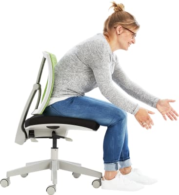 Eine Frau mit Brille und grauem Pullover sitzt nach vorne gebeugt auf einem Bürostuhl und streckt die Arme vor sich aus. Sie trägt Jeans und weiße Turnschuhe auf weißem Grund.