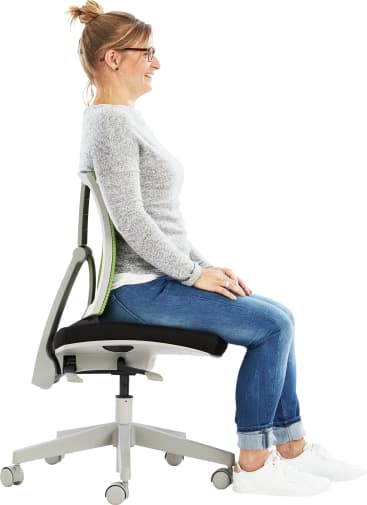 Eine Frau mit Brille und grauem Oberteil sitzt aufrecht auf einem Bürostuhl, die Hände auf den Oberschenkeln, die Füße flach auf dem Boden, den Rücken gerade an die grün akzentuierte Lehne gelehnt.