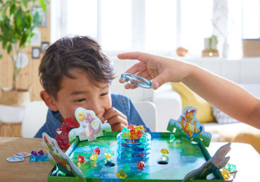 Ein kleines Kind beobachtet, wie eine andere Hand einen durchsichtigen Plastikstein auf ein buntes Brettspiel legt, das von kleinen farbigen Edelsteinen und illustrierten Tierausschnitten auf einem Tisch in einem hellen Raum umgeben ist.