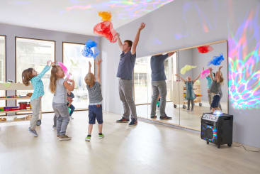 Ein Erwachsener und fünf kleine Kinder stehen in einem hellen Raum und werfen bunte Tücher in die Luft. An der Wand sind Lichtmuster zu sehen, und auf dem Boden steht ein Lautsprecher. Im Hintergrund sind ein großer Spiegel und Musikinstrumente zu sehen.