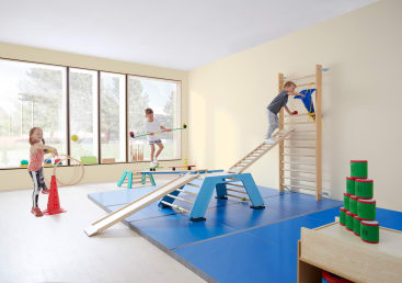 Drei Kinder spielen in einer hellen Turnhalle mit weichen blauen Matten, Schwebebalken und Klettergeräten. Ein Kind klettert auf eine Leiter, ein anderes balanciert auf einem Balken, und ein drittes benutzt Ringe. Ein Stapel grüner und roter Becher steht auf einem Tisch.