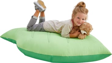 Ein kleines Kind liegt auf einem großen, hellgrünen Kissen, hält einen braunen Plüschhund und lächelt in die Kamera. Das Kind trägt Jeans, ein helles Hemd und Turnschuhe.