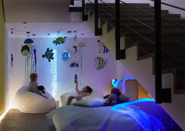 Zwei Kinder entspannen sich auf weißen Sitzsäcken in einem schwach beleuchteten Raum unter einer Treppe, umgeben von blauen und lilafarbenen sensorischen Lichtern und Wanddekorationen zum Thema Unterwasser, darunter Fische und Quallen.