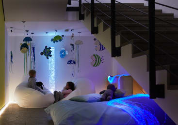 Zwei Kinder sitzen auf weißen Sitzsäcken unter einer Treppe in einem sanft beleuchteten Raum mit einem Unterwasserthema, mit Wanddekorationen von Meerestieren und beleuchteten Sinneslichtern. Ein Plüschtier liegt auf einem Bett in der Nähe.