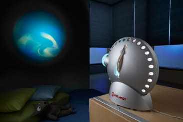 Ein Mathmos Space Projector steht auf einem Tisch und projiziert ein blaues, wirbelndes Lichtmuster auf eine dunkle Wand. Im Hintergrund sind Kissen, ein Plüschtier und Fenster mit teilweise geschlossenen Jalousien zu sehen.