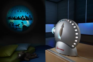 Ein Mathmos-Projektor steht auf einem Tisch und projiziert eine skurrile Nachtszene mit einer Stadtsilhouette, Sternen und einer Katze an eine Wand. Der Raum ist schwach beleuchtet, die Jalousien sind geschlossen und ein Kuscheltier liegt auf einem Kissen in der Nähe.