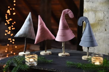 Vier Stoffornamente in Form von Bäumen und Hüten stehen auf einem Tisch mit kleinen Zweigen als Stämmen. Im Hintergrund sind Tannenzweige, zwei Glasvotivkerzen und verschwommene Lichterketten zu sehen.