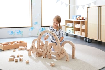 Ein kleines Kind sitzt auf einem runden beigen Teppich und baut mit Holzklötzen ein großes Bauwerk in einem hellen, aufgeräumten Raum mit Regalen, Kissen und Wanddekorationen.