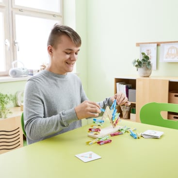 Ein junger Mann sitzt an einem grünen Tisch und befestigt bunte Wäscheklammern an einem hölzernen Dreieck, auf dem Tisch in einem hellen, aufgeräumten Raum liegen weitere Wäscheklammern und Karten.