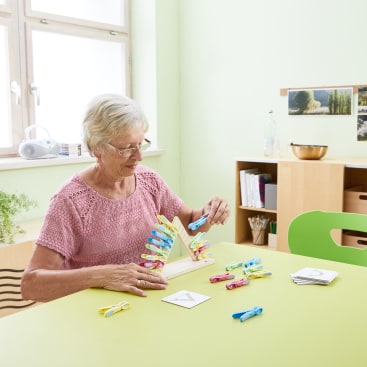 Eine ältere Frau mit kurzen grauen Haaren und Brille sitzt an einem grünen Tisch und befestigt bunte Wäscheklammern an einem Holzständer. Auf dem Tisch in einem hellen, aufgeräumten Raum liegen Karten und weitere Wäscheklammern.