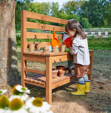 Ein kleines Kind in einem grauen Hemd, braunen Shorts und gelben Regenstiefeln steht an einer hölzernen Spielbank im Freien und arrangiert kleine Töpfe und Küchenutensilien in einer Gartenlandschaft.
