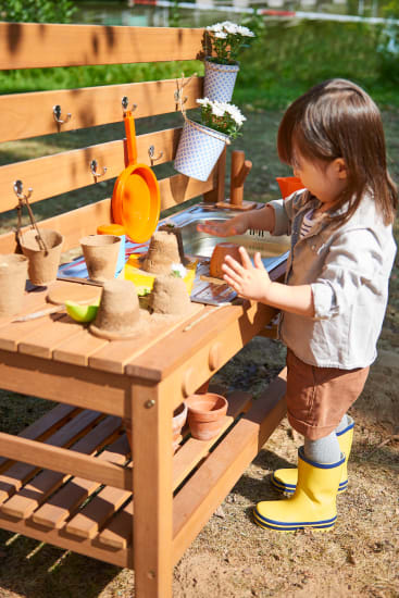 Ein kleines Kind in gelben Regenstiefeln steht an einem hölzernen Spieltisch im Freien und baut Sandstrukturen mit kleinen Töpfen und Küchenutensilien. Pflanzen und Blumen hängen an der Bank im Hintergrund.
