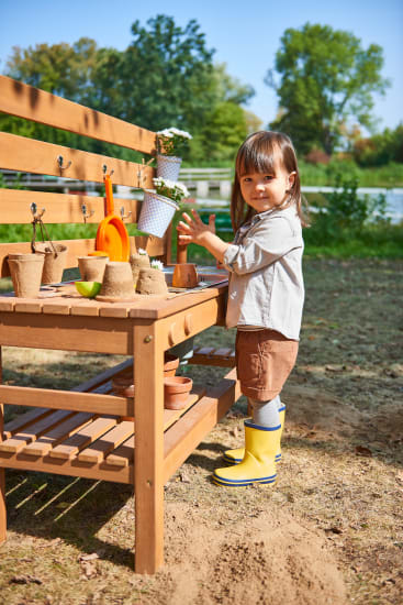 Ein kleines Kind in gelben Regenstiefeln steht an einem sonnigen Tag an einer hölzernen Werkbank im Freien mit Erde, Töpfen und Gartenwerkzeugen. Im Hintergrund sind Bäume und Grünflächen zu sehen.