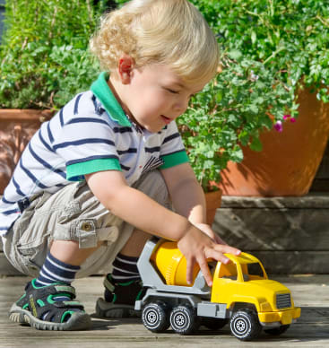 Ein kleines Kind mit blondem Haar, das ein gestreiftes Polohemd und Shorts trägt, hockt im Freien und spielt mit einem gelben Spielzeug-Zementmischwagen auf einer Holzfläche. Im Hintergrund sind Topfpflanzen zu sehen.