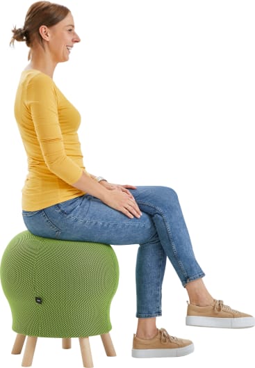 Eine Frau in einem gelben Oberteil und blauen Jeans sitzt seitlich auf einem grünen, stoffbezogenen Gymnastikstuhl mit Holzbeinen, lächelt und blickt nach vorn.