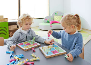 Zwei kleine Kinder sitzen an einem Tisch und spielen mit Reißzwecken und bunten Wäscheklammern in einem hellen, modernen Raum mit einem grün-weißen Sofa und einem großen Fenster im Hintergrund.