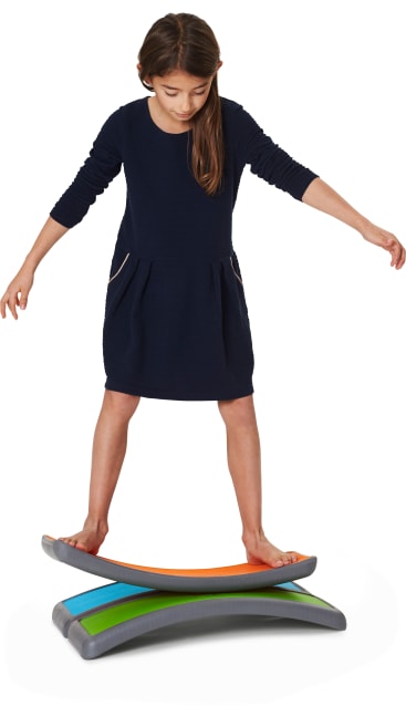 Ein junges Mädchen in einem marineblauen Kleid steht barfuß auf einem orangefarbenen Balancierbrett, das auf zwei anderen Balancierbrettern, einem blauen und einem grünen, aufgestapelt ist. Sie balanciert mit ausgestreckten Armen, um sich abzustützen.