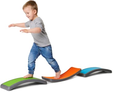 Ein kleines Kind läuft barfuß auf bunten, geschwungenen Balancierbalken. Es trägt ein graues, langärmeliges Hemd und blaue Jeans und streckt die Arme aus, um vor einem weißen Hintergrund das Gleichgewicht zu halten.