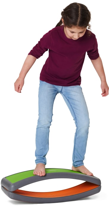 Ein Kind mit langen braunen Haaren, einem kastanienbraunen Hemd und einer blauen Jeans steht barfuß auf einem gebogenen grauen Balancierbrett mit grünen und orangefarbenen Flächen. Das Kind schaut nach unten und balanciert mit leicht ausgestreckten Armen.
