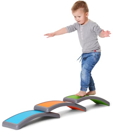 Ein kleines Kind mit blondem Haar, das ein graues Hemd und eine blaue Jeans trägt, balanciert barfuß auf geschwungenen, bunten Trittsteinen, die in einer Reihe auf einem weißen Hintergrund angeordnet sind.