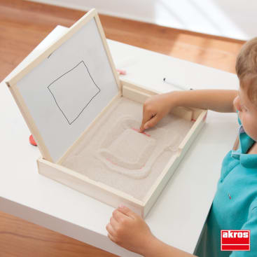 Ein Kind sitzt an einem weißen Tisch und zeichnet mit seinem Finger eine Form in einen mit Sand gefüllten Kasten. Auf dem Deckel der Schachtel ist ein Whiteboard angebracht, auf dem eine rechteckige Form gezeichnet ist. In der rechten unteren Ecke ist das Akros-Logo zu sehen.
